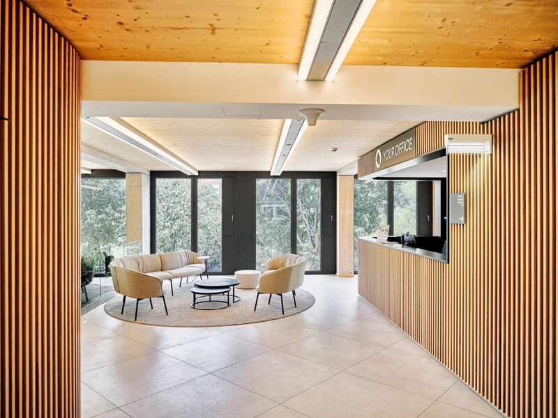 Moderne Büro-Lobby und Empfangstheke mit Holz-Lamellenwänden, gemütlicher Lounge-Sitzgruppe und großen Fenstern mit Blick ins Grüne.