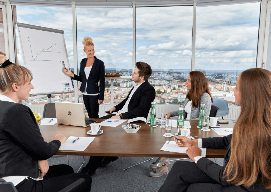 Ein divers besetztes Business-Team arbeitet zusammen und folgt einer Präsentation in einem Hochhaus-Büro mit Blick auf die Stadt.