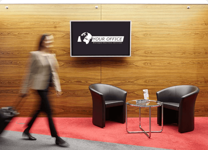 A modern office lobby with a company logo on the screen, a waiting area with black armchairs, and a person walking with luggage.