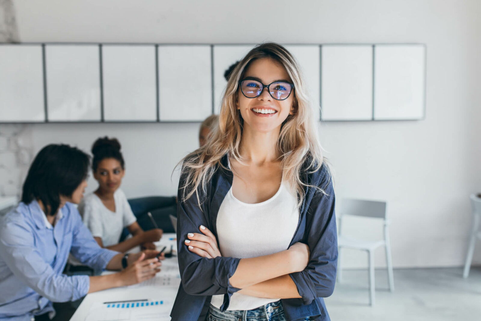 Lächelnte blonde Sekretärin mit trendiger Brille und verschränkten Armen in einem modernen Büro während eines Meetings