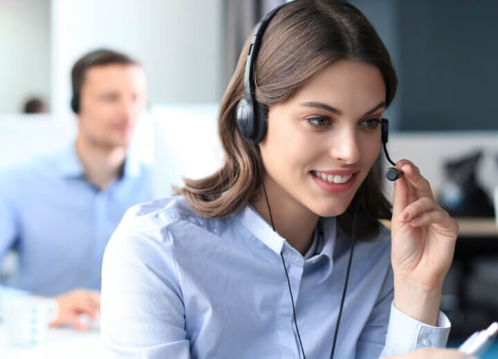 A smiling customer service agent wearing a headset while working in an office with a colleague in the background.