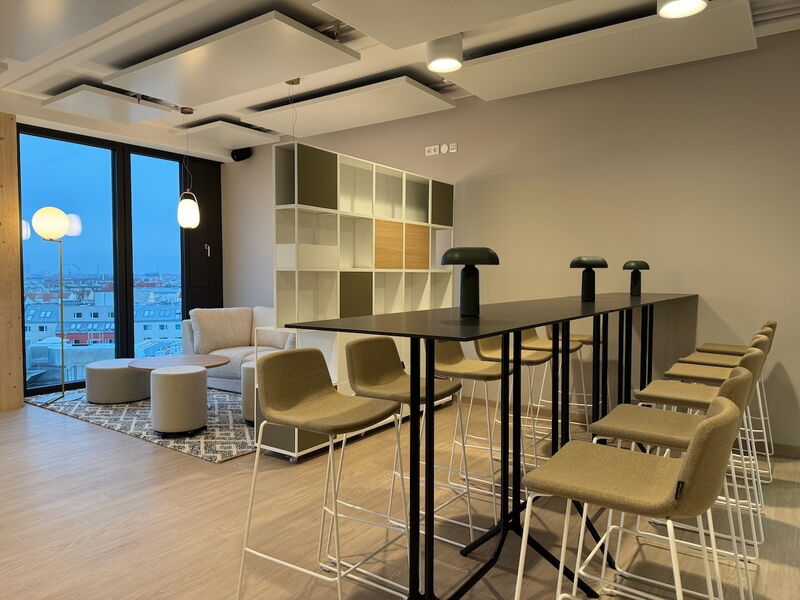 Geräumiges, modernes Büro mit einem hohen Stehtisch mit grünen Hockern, einem gemütlichen Loungebereich mit Sofa und einem großen Fenster mit Blick auf die Stadt bei Dämmerung.