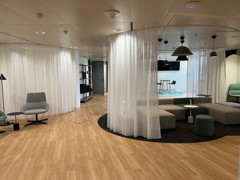 Modernes Großraumbüro mit modularen Sofas, die durch weiße, transparente Vorhänge abgetrennt sind, Holzboden und zeitgemäßen Sitzmöbeln.