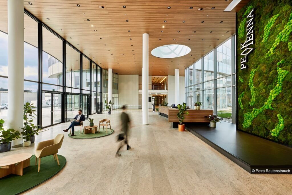 Spacious and modern office lobby interior at Peak Vienna featuring a large vertical green moss wall, wooden ceiling, and large glass windows.
