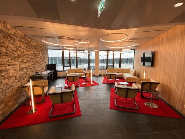 Spacious modern office lounge area featuring comfortable beige armchairs, sofas, red carpets, a feature stone wall, and stylish circular pendant lighting.
