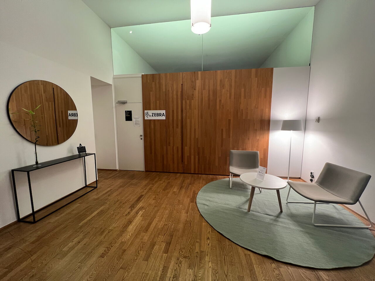 Heller Wartebereich im Büro mit Holzpaneelen, rundem Spiegel, Sesseln und einem Couchtisch auf einem grünen Teppich.