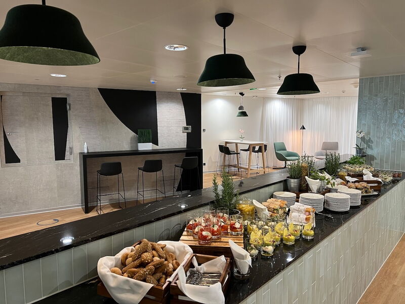 Ein gut gefülltes Buffet in einem modernen Speisesaal mit frischem Gebäck, kleinen Dessertgläsern, Salaten und Stapeln von weißen Tellern.