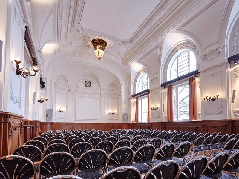 Geräumiger historischer Saal mit verzierten weißen Stuckdecken, Bogenfenstern, Holzvertäfelung und Reihen von leeren Stühlen.