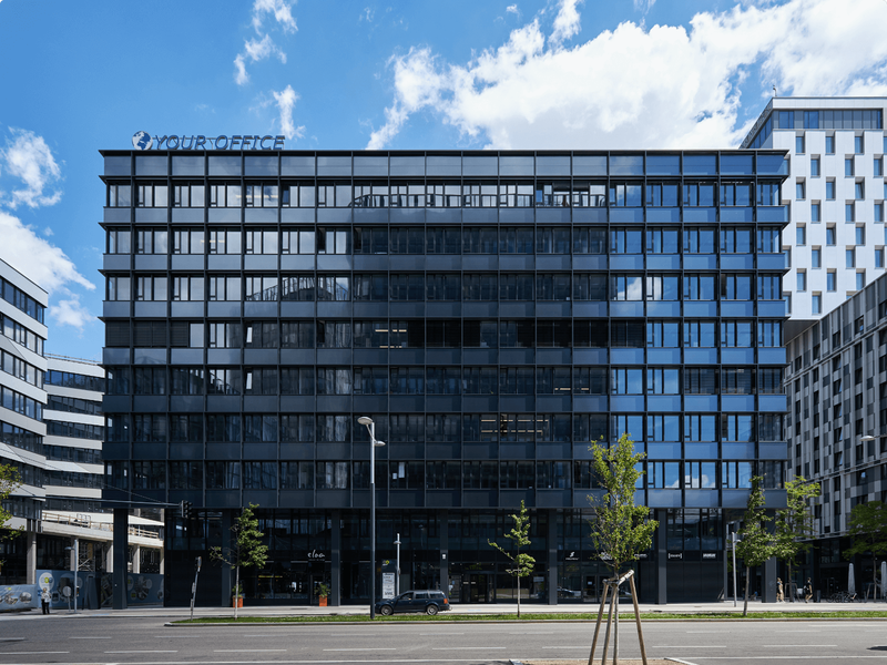 Außenansicht eines modernen, dunklen Glas-Bürogebäudes mit Your Office Schriftzug vor blauem Himmel mit Wolken