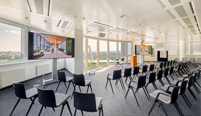 Heller, moderner Konferenz- und Veranstaltungsraum im Peak Vienna mit schwarzen Stuhlreihen, Präsentationsbildschirmen und Panoramablick über die Stadt und den Fluss.