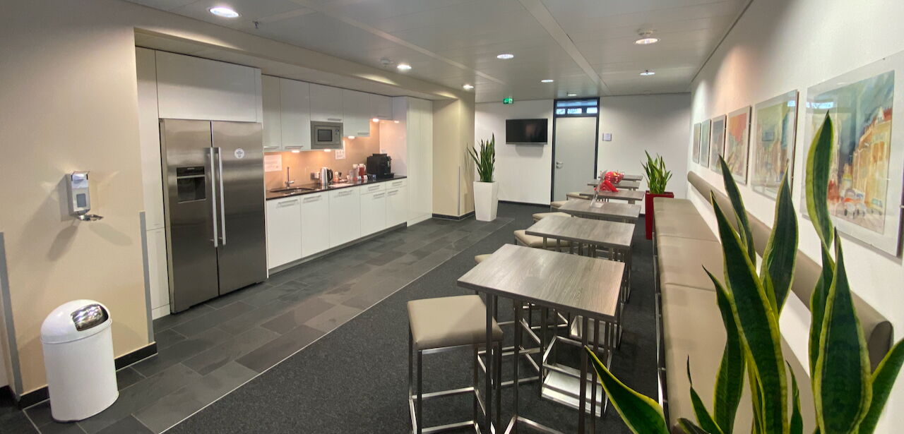 Schicke, moderne Büro-Teeküche mit grauen Küchenmöbeln, einer Kaffeemaschine und hohen Stehtischen mit Stühlen auf Fliesenboden.
