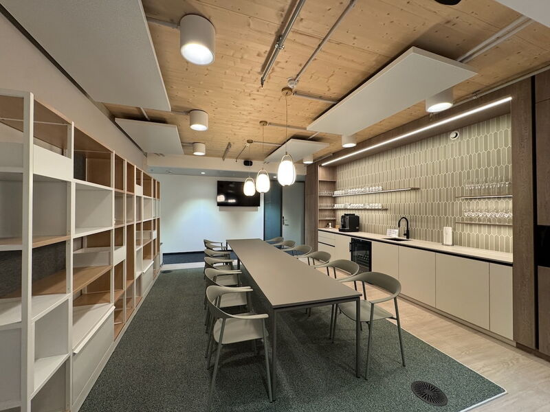 Ein moderner Büro-Pausenraum mit langem Konferenztisch, Teeküche mit Kaffeemaschine und hölzerner Akustikdecke.