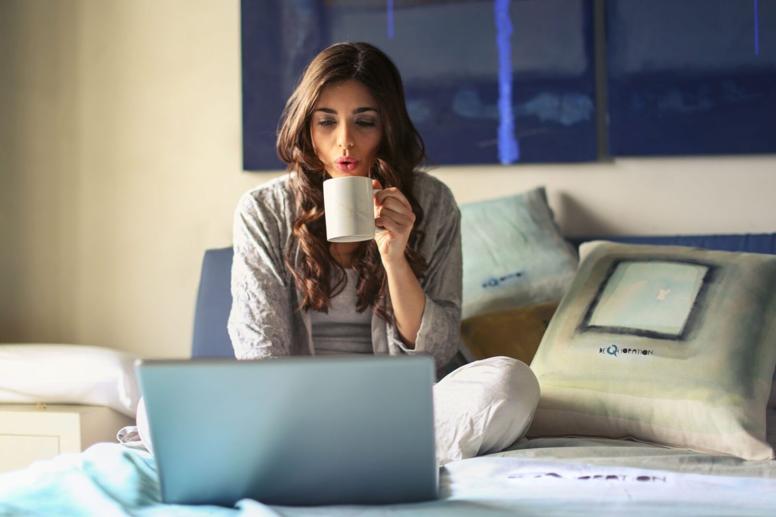 Junge Frau trinkt Kaffee aus einer weißen Tasse und arbeitet mit einem Laptop im Bett
