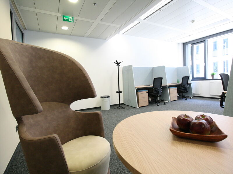 Moderne Büro-Lounge mit einem braunen Ohrensessel, einem Holztisch mit Äpfeln und abgetrennten Arbeitsplätzen im Hintergrund im Euro Plaza.