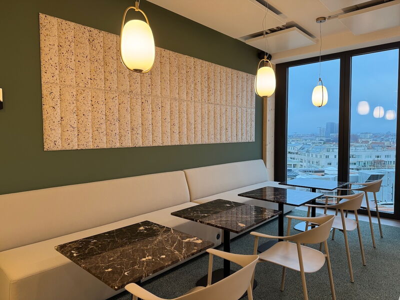 Moderne Büro-Lounge mit einem langen beigen Sofa, dunklen Marmortischen, Hängeleuchten, Akustik-Wandpaneelen und einem großen Fenster mit Blick auf die Stadt.