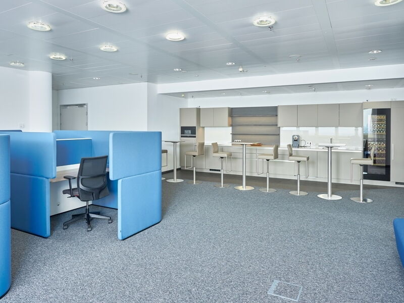 Modern office interior featuring blue acoustic cubicles, workstations, and a fully equipped kitchen and breakroom area with bar stools.