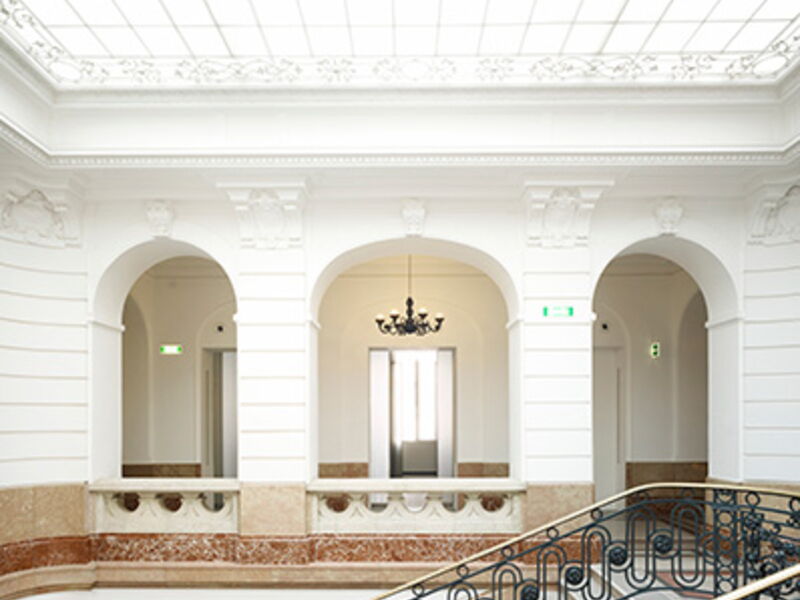 Helles historisches Foyer mit weißen Rundbögen, verzierten Säulen, Glasdach und klassischem Kronleuchter