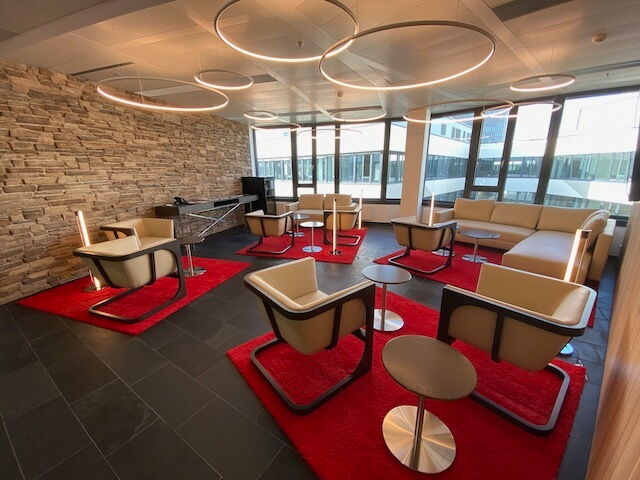 Bright modern office lounge area featuring beige armchairs, red rugs, circular ring lighting, and a stone accent wall.