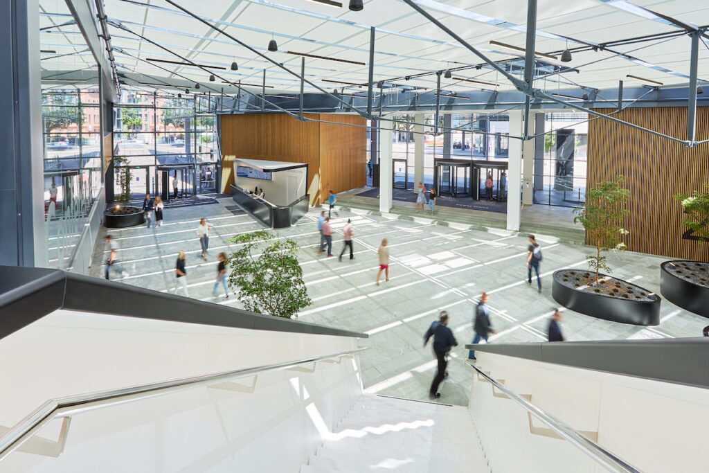 Spacious modern corporate office lobby interior featuring a glass atrium roof, wooden wall accents, indoor trees, and people walking across the stone floor.