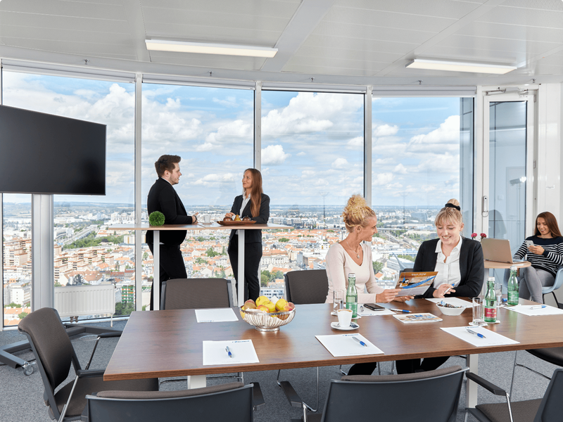 Ein Business-Team arbeitet in einem modernen Hochhaus-Konferenzraum mit großen Fenstern und Blick über die Skyline zusammen.