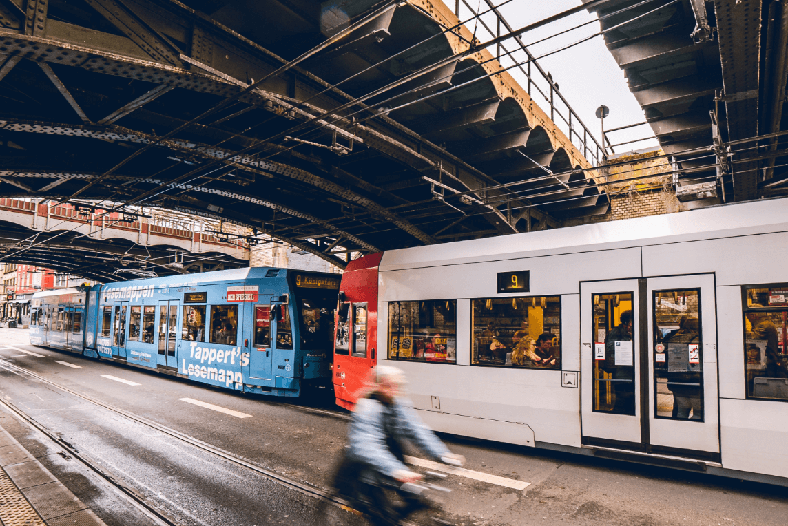 Zwei moderne Straßenbahnen, eine blaue und eine weiße, fahren unter einer historischen Hochbahnbrücke in einer Großstadt