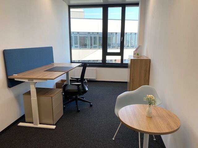 Modernes Bürointerieur mit einem höhenverstellbaren Schreibtisch in Holzoptik, blauer Akustikwand, Bürostuhl, kleinem runden Besprechungstisch und großem Fenster.