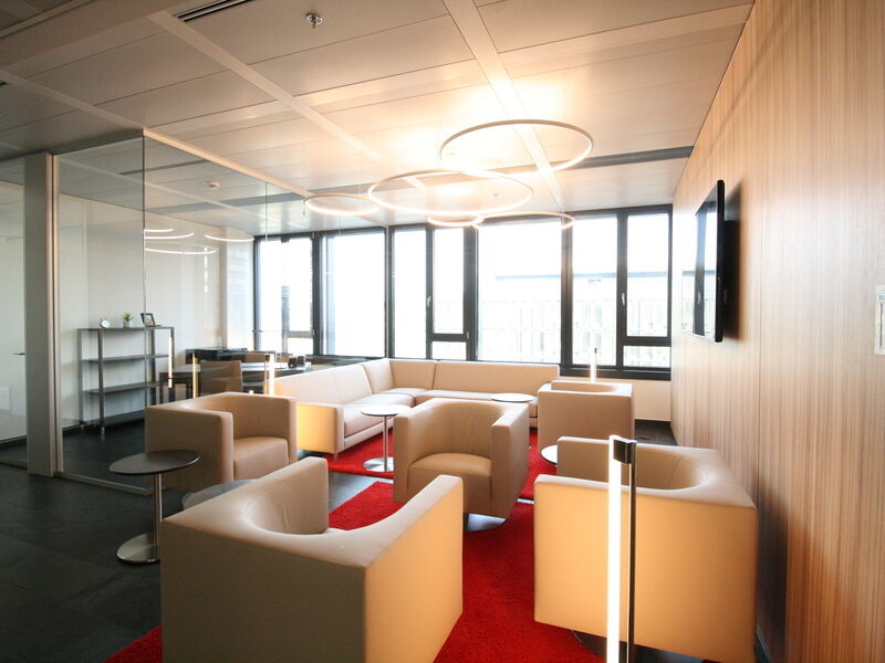 Großzügige, moderne Büro-Lounge mit beigen Sesseln, einem großen Ecksofa, rotem Teppich und auffälligen runden Pendelleuchten.