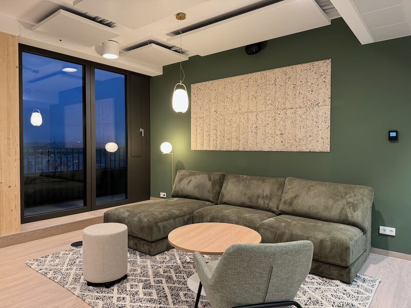 Gemütliche Büro-Lounge mit großem grünen Cord-Ecksofa, einem Holztisch und schalldämmenden Paneelen an einer grünen Wand.