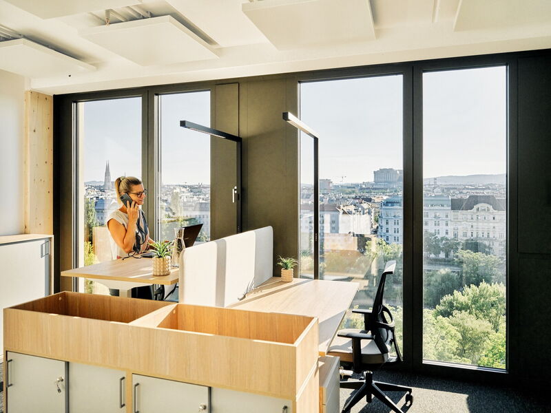 Geschäftsfrau telefoniert an einem ergonomischen Stehschreibtisch in einem modernen Büro mit großen Panoramafenstern und Blick auf die Skyline