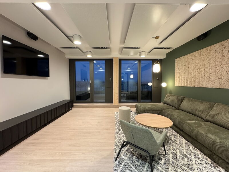 Geräumiger, moderner Büro-Lounge-Bereich mit großem grünen Cord-Sofa, Akustikpaneelen an Decke und Wand, Fernseher und großen Fenstern in der Dämmerung.