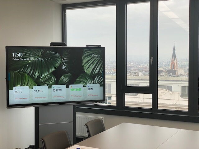 A modern meeting room featuring a large interactive digital display showing room climate data and a view of Vienna through the window.