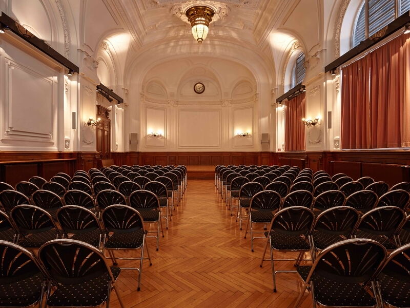Ein geräumiger, eleganter Saal mit klassischen architektonischen Details, einem Gewölbedecke mit Kronleuchter, Holzvertäfelung und ordentlich angeordneten Reihen schwarzer Stühle mit Blick auf eine Bühne.