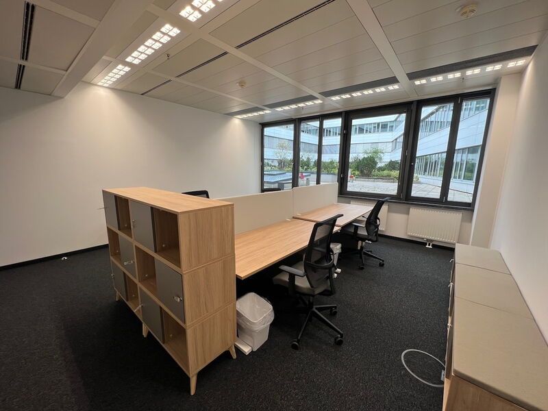 Helles und modernes, leeres Büro mit Holztischen, ergonomischen Stühlen, einem Raumteiler-Regal und großen Fenstern mit Blick auf einen Innenhof.