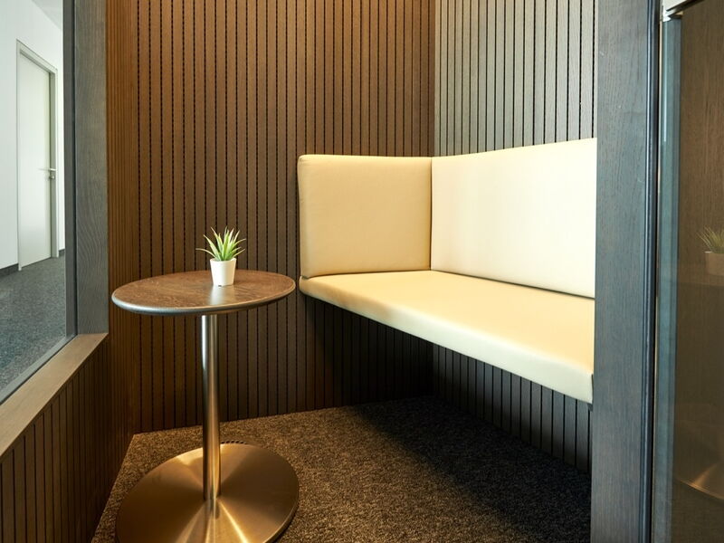 Innenansicht einer ruhigen Büro-Besprechungszelle mit cremefarbener Sitzbank, Holz-Akustikpaneelen und einem kleinen runden Tisch.