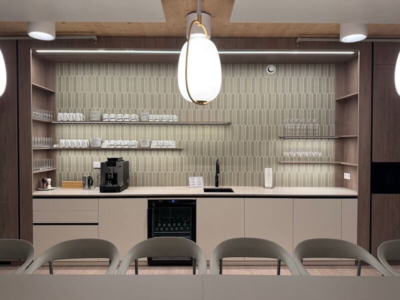 Stilvolle Büroküche mit beigen Fliesen im Picket-Design, Regalen aus Holz, Kaffeevollautomat, Weinkühlschrank und moderner Hängeleuchte.