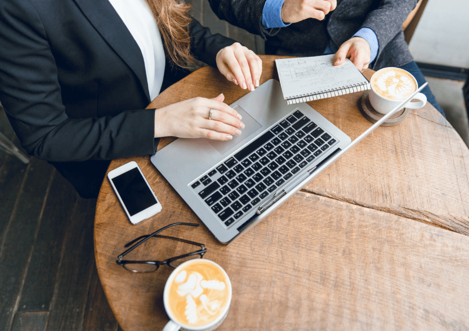 Two business professionals collaborating in a coffee shop with laptops, notebooks, and coffee cups on a wooden table.