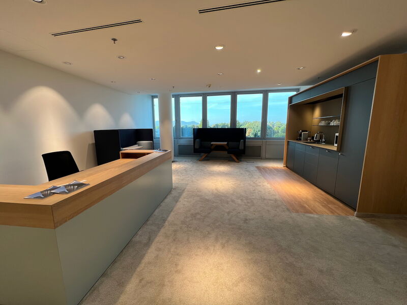 Geräumiges, modernes Büro mit einer Empfangstheke im Vordergrund, einer integrierten Teeküche rechts und gemütlichen Sitzbereichen an den großen Fenstern.