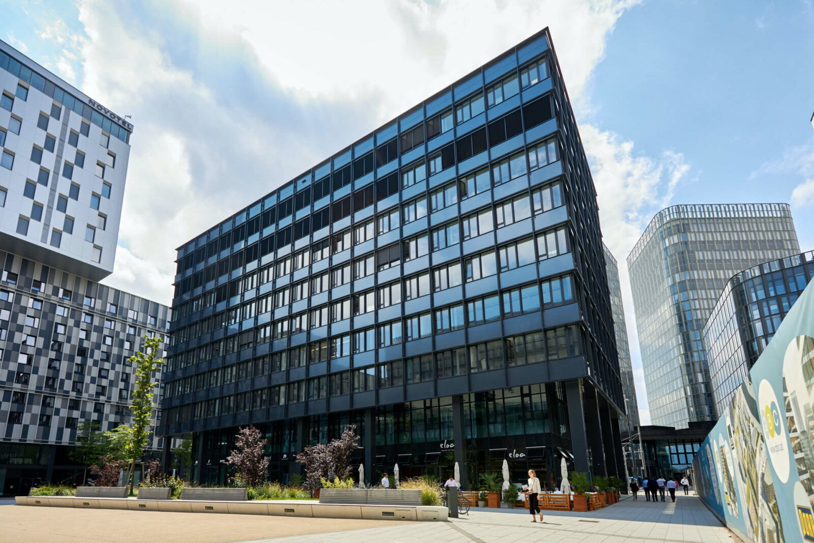 Außenansicht eines modernen, dunklen Glas-Bürogebäudes mit umliegenden Wolkenkratzern und Platz in urbaner Lage