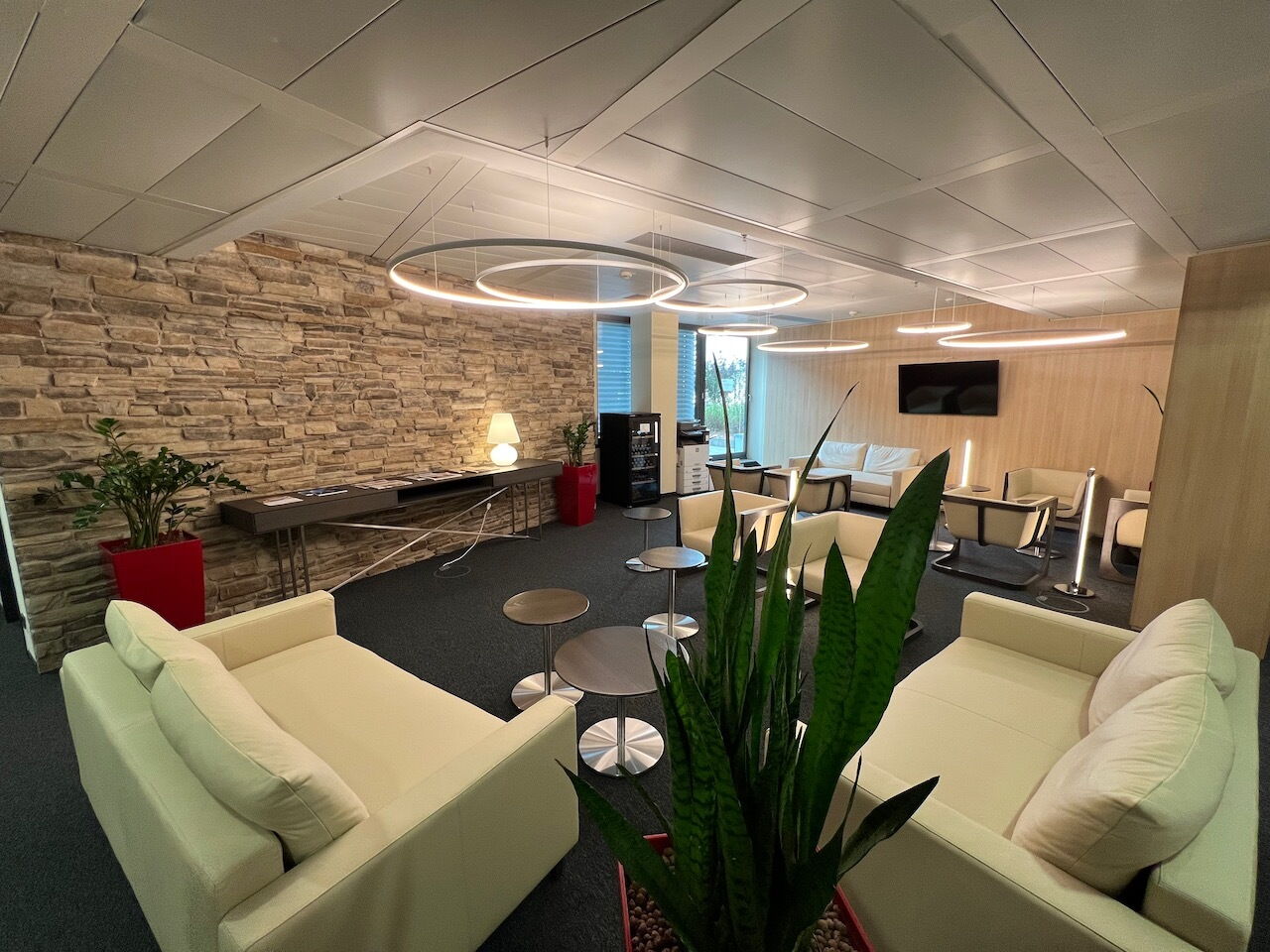 Geräumige moderne Büro-Lounge mit bequemen weißen Sofas, einer rustikalen Steinakzentwand und runden Pendelleuchten.