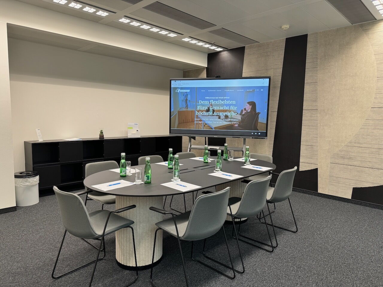 Moderner Konferenzraum mit einem ovalen Besprechungstisch, grauen Stühlen, Wasserflaschen und einem großen Präsentationsbildschirm an der Wand.
