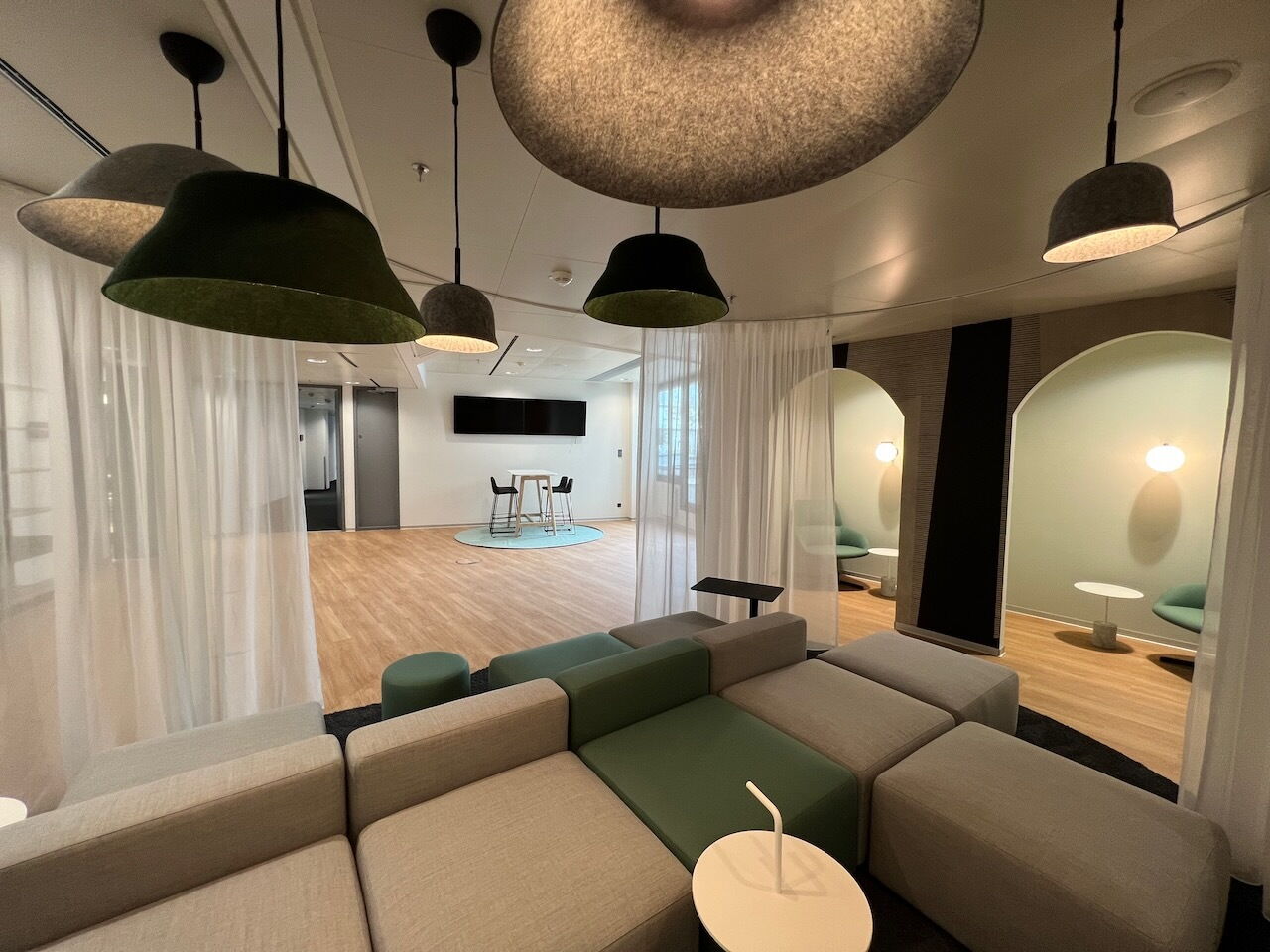Großzügige, moderne Büro-Lounge mit modularen grau-grünen Sofas, Akustik-Filz-Pendelleuchten und halbrunden Besprechungsnischen mit weißen Vorhängen.