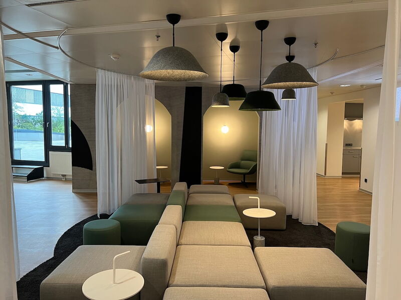 Ein moderner Lounge-Bereich im Büro mit weißen Vorhängen, einem grünen Sessel, einem grauen Sessel und einem Sofa.