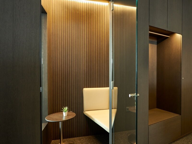 Moderne Büro-Telefonzelle mit Holzlamellen-Akustikwandpaneelen, cremefarbener Eckbank, kleinem Beistelltisch und Glastür neben integrierter Garderobe.