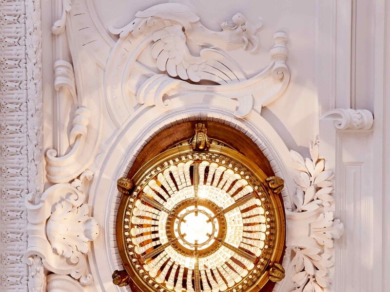 Nahaufnahme eines prachtvollen goldenen Kronleuchters, eingefasst in filigranen weißen Barock-Stuck an einer historischen Decke.