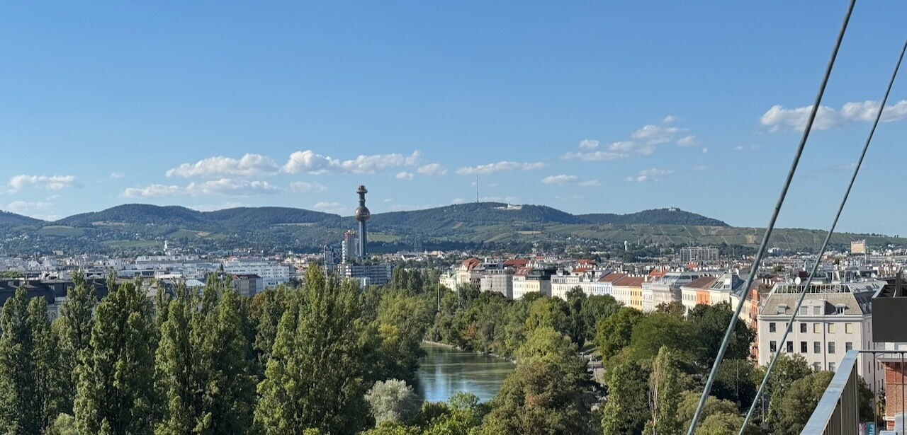 Panoramablick auf das Wiener Stadtbild mit dem Donaukanal, grünen Bäumen und den Hügeln im Hintergrund unter blauen Himmel.