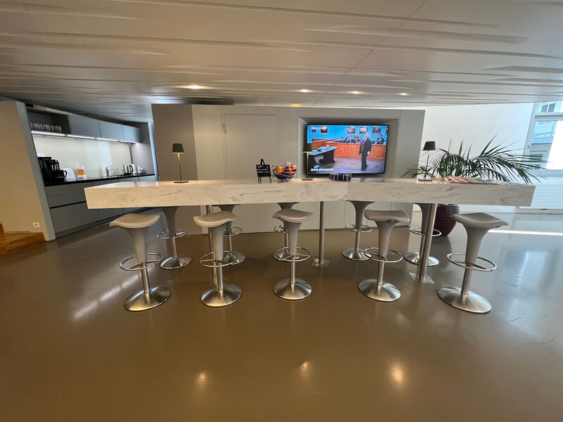 Moderne Büro-Pausenzone mit einer langen Marmor-Bar, eleganten grauen Barhockern, einer Teeküche im Hintergrund und einem Flachbildschirm.