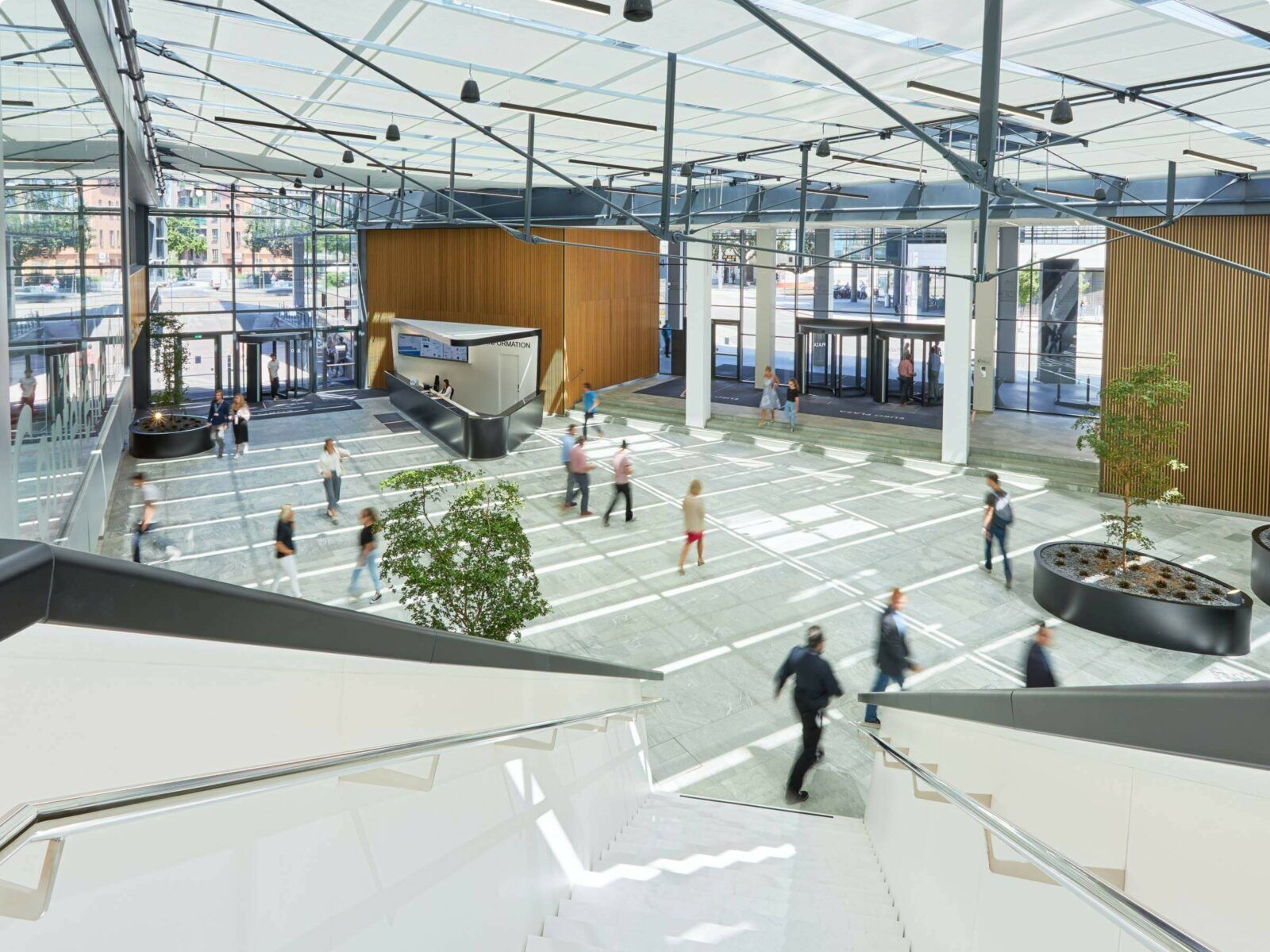 Großzügiges, modernes Büro-Foyer mit hohem Glasdach, Holzelementen und gehendem Personal, aus der Vogelperspektive betrachtet.