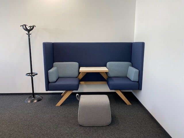 Blaue Akustik-Sitzbox fürs Büro mit hoher Rückenlehne, integriertem Holztisch und grauen Hocker in einem modernen Meetingraum