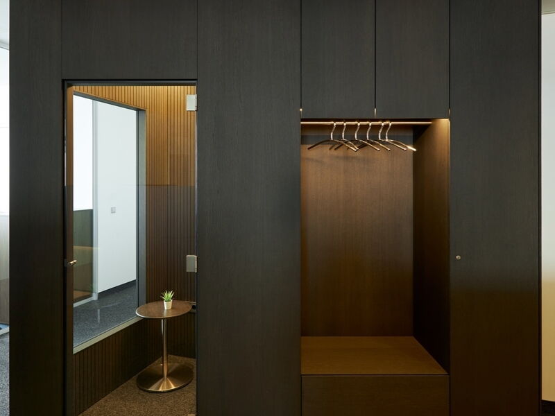 Zeitgenössische dunkle Holzwand mit integrierter Garderobe, beleuchteten Kleiderbügeln und einer angrenzenden Glastür, die zu einem modernen Büro-Besprechungsraum mit einem kleinen Tisch führt.