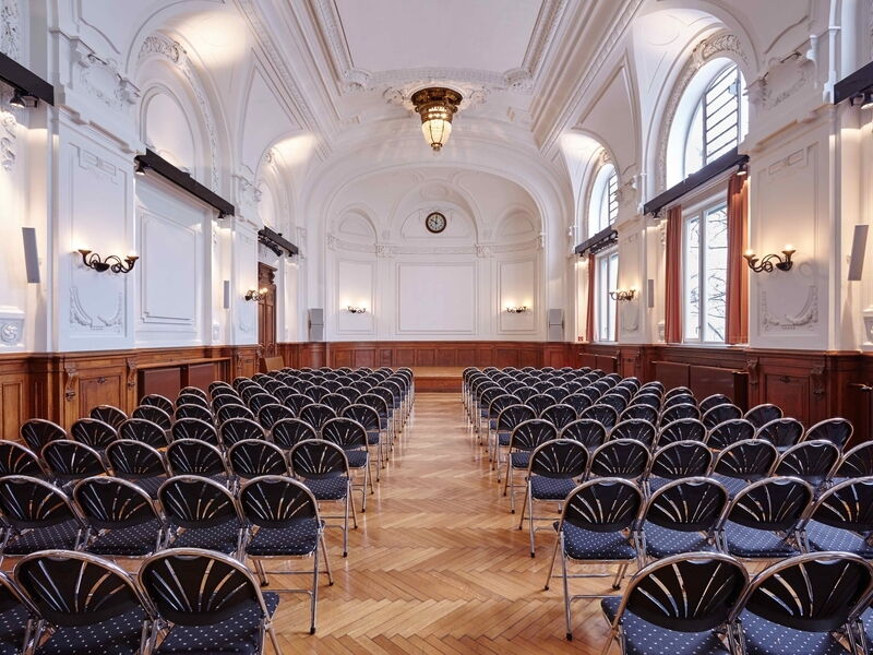 Geräumiger Veranstaltungssaal mit verzierten weißen Wänden, Holzvertäfelung, Bogenfenstern und Reihen dunkler Stühle für eine Präsentation.